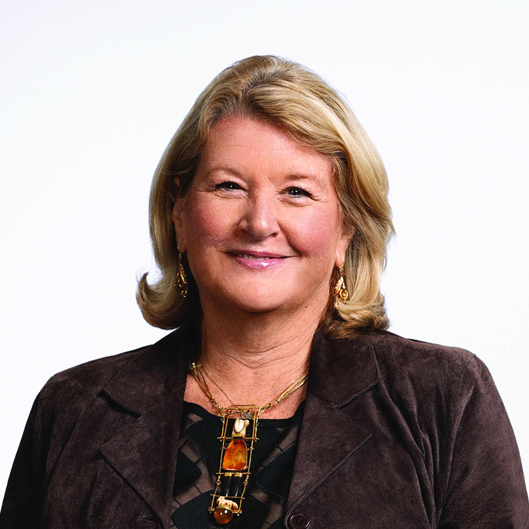 A middle-aged woman with blonde hair wears a brown jacket, patterned top, gold earrings, and a large gold necklace, smiling against a plain light background.