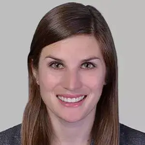 A woman with straight brown hair, fair skin, and brown eyes is smiling at the camera against a plain light gray background.