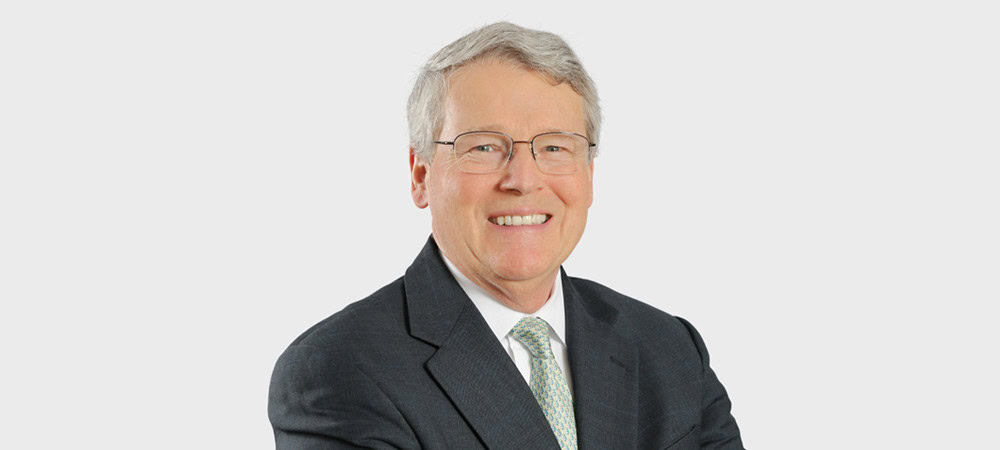 Older man with gray hair and glasses wearing a dark suit, light shirt, and patterned tie, smiling against a plain light background.