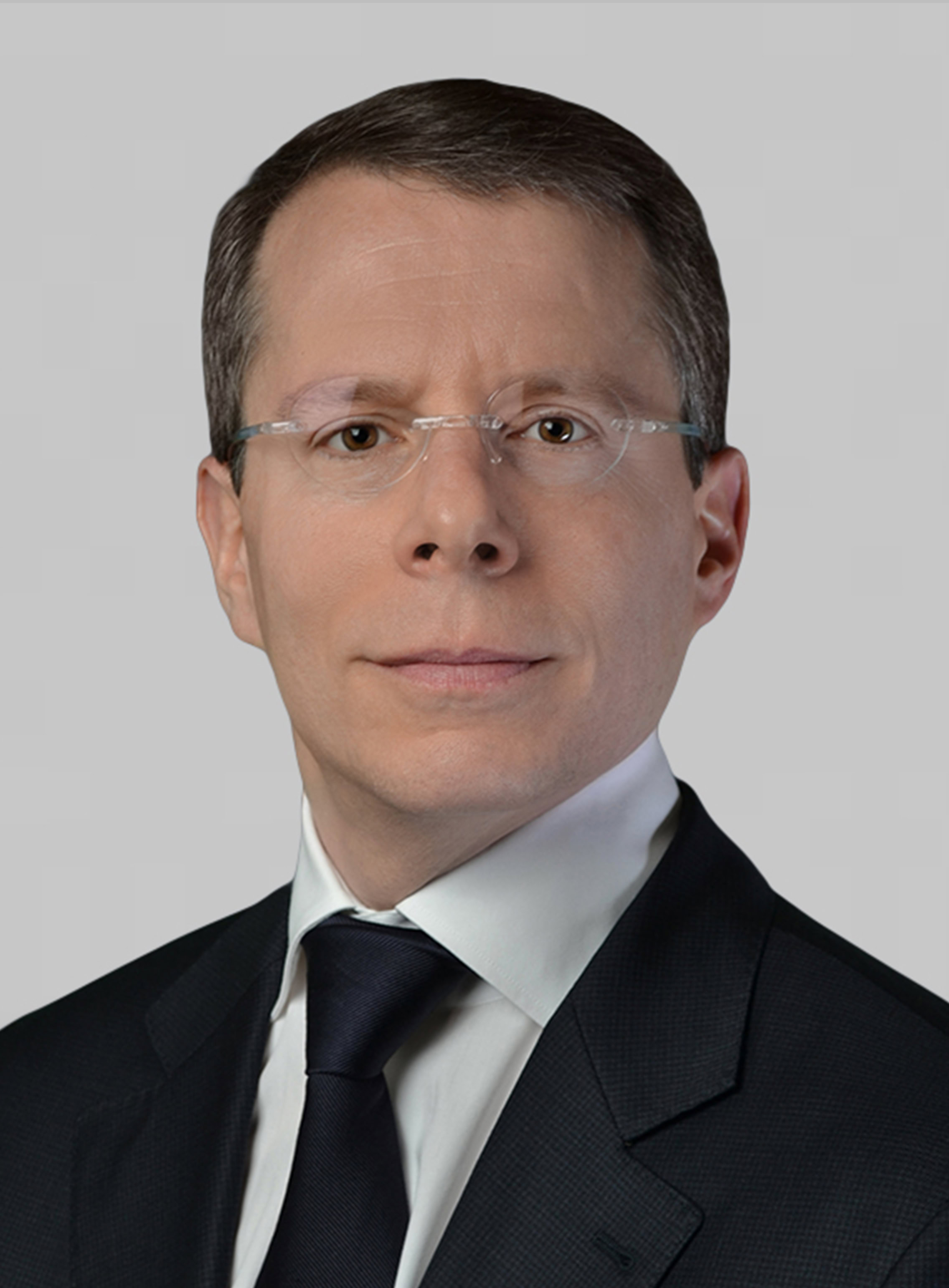 A man wearing glasses, a dark suit, white shirt, and dark tie, facing forward against a plain light gray background.