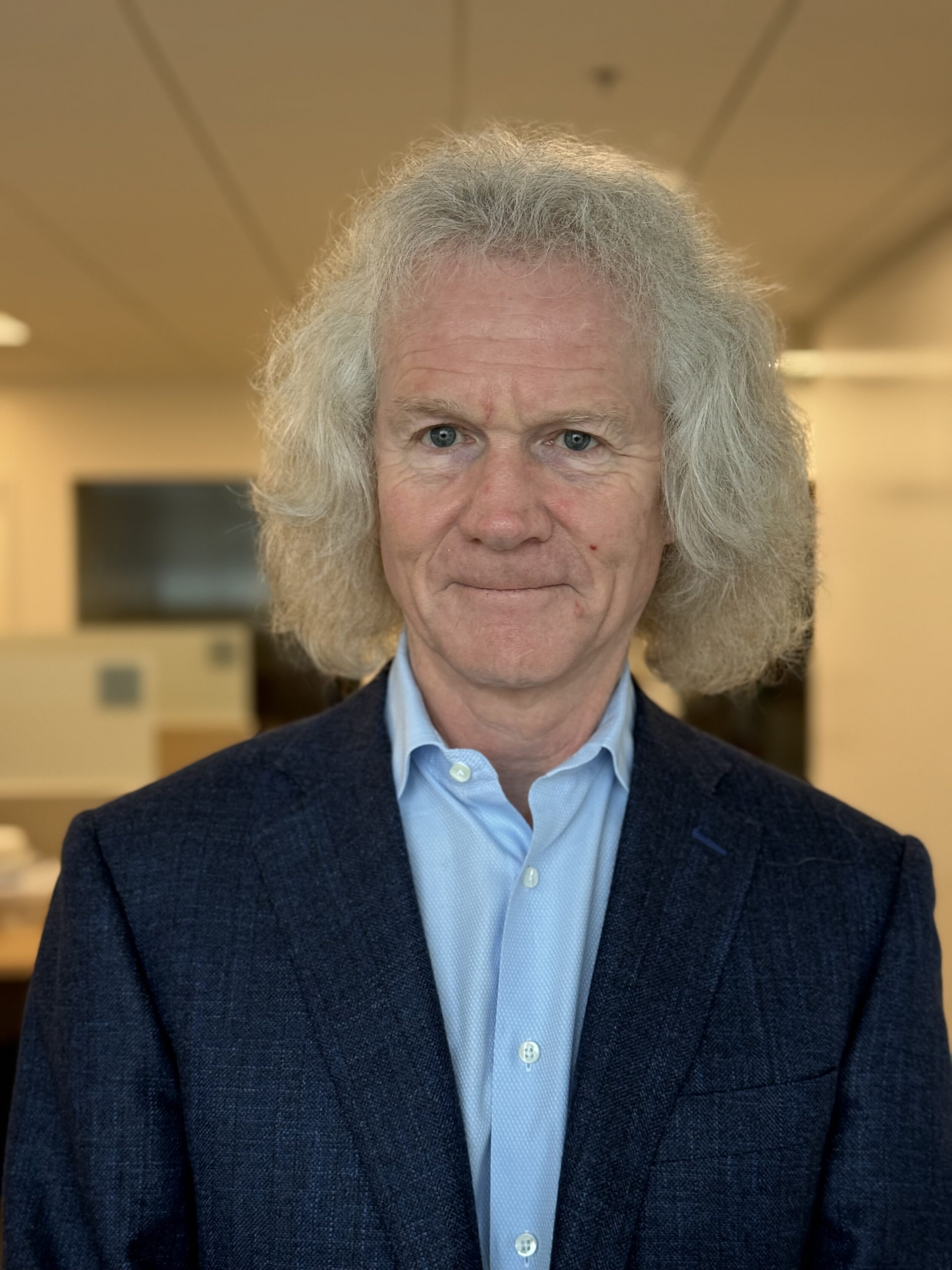 A man with white curly hair wearing a blue dress shirt and dark blazer stands indoors, facing the camera with a neutral expression.