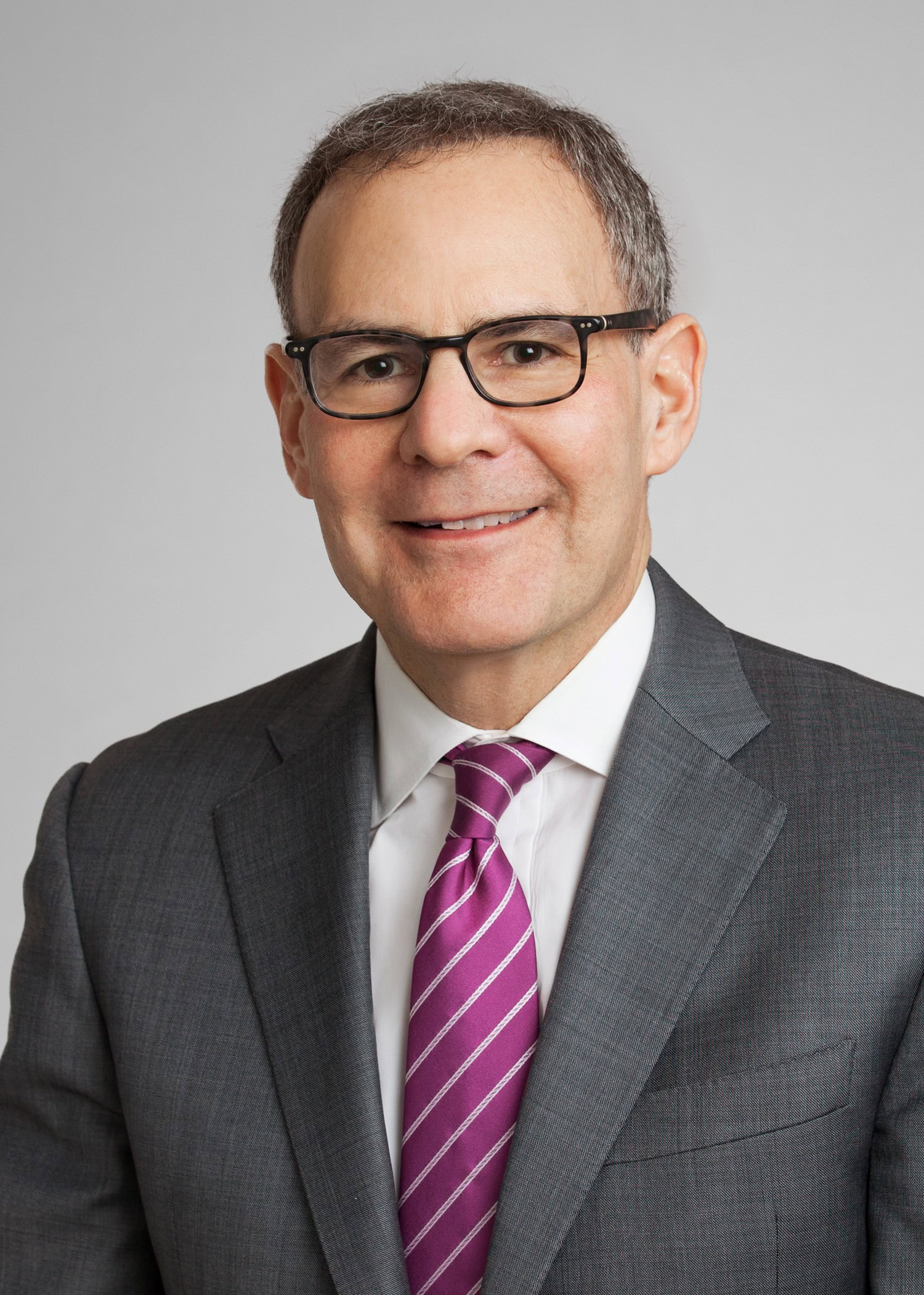 A man wearing glasses, a gray suit, white shirt, and purple striped tie, posed against a plain light background.