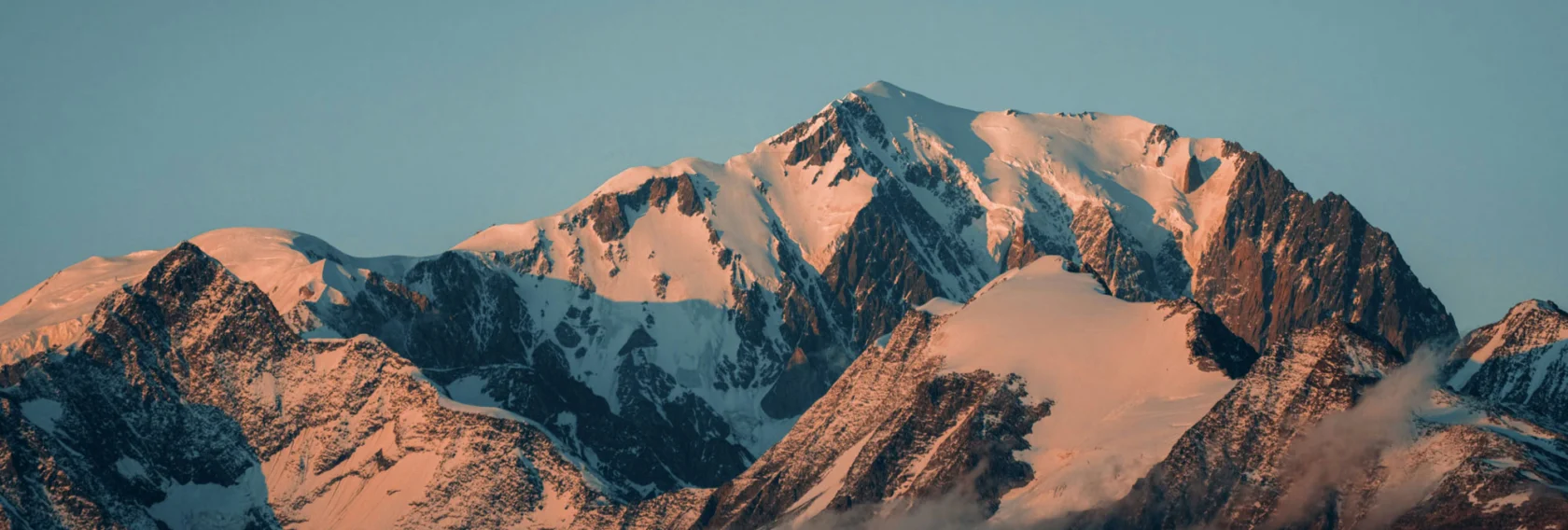 Snow-capped mountain peaks with rocky faces lit by warm sunrise light against a clear blue sky.