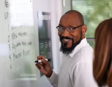 A man in glasses and a white shirt writes on a whiteboard while smiling, with a woman standing nearby.