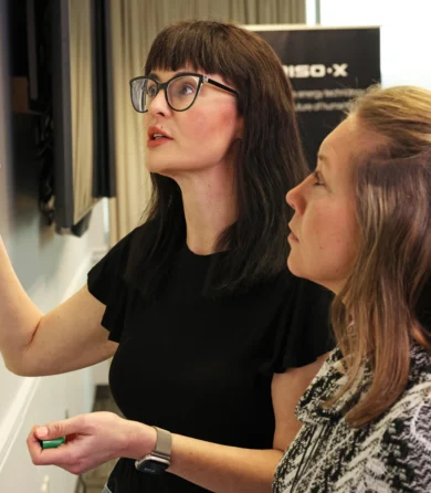 Two women stand at a whiteboard, one writing with a green marker while the other watches, in a modern office setting.