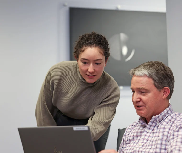A woman stands next to a seated man, both looking at a laptop screen in an office setting.