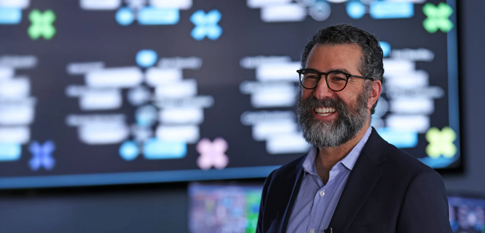 A man with glasses and a beard smiles while standing in front of a large digital screen displaying blurred diagrams and information.