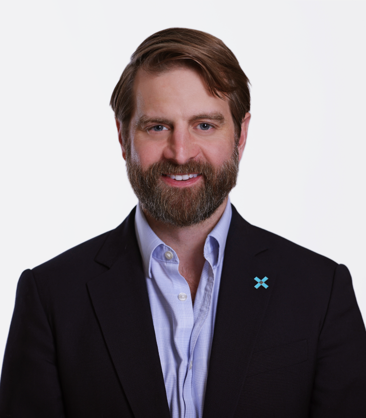 A man with brown hair and a beard wearing a navy blazer and light blue shirt, standing against a plain white background.