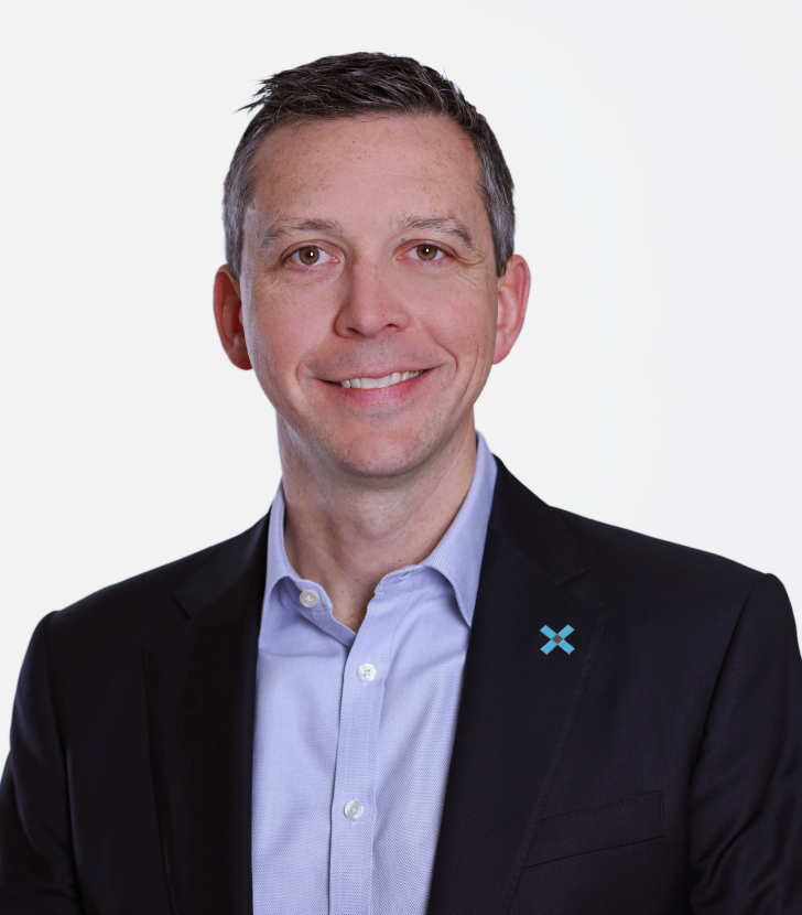 A man in a dark suit jacket and light blue shirt, smiling, against a plain white background. A blue “X” pin is on his left lapel.
