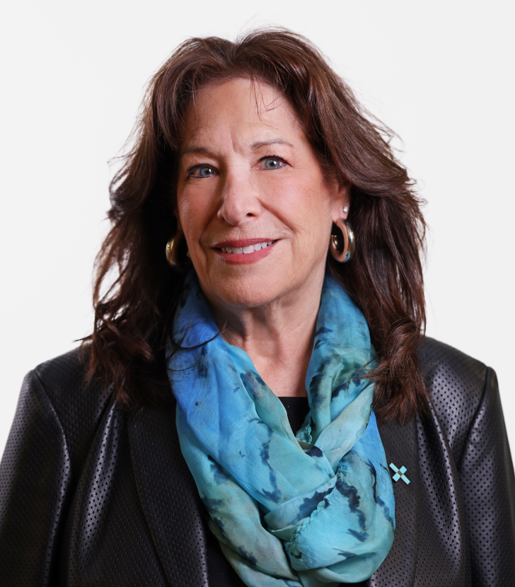 Woman with medium-length brown hair, wearing a black jacket, large hoop earrings, and a blue patterned scarf, standing against a plain white background.