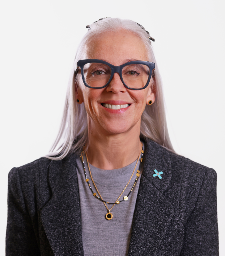 A woman with long gray hair and large glasses, wearing a gray blazer, gray top, layered necklaces, and a blue cross pin, smiles at the camera against a plain background.