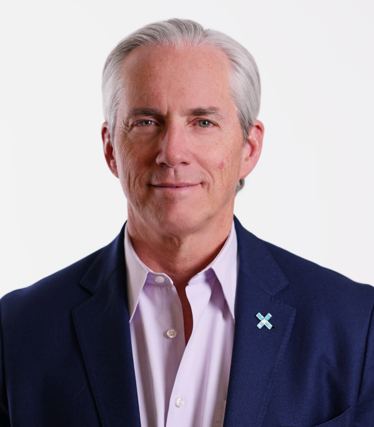 An older man with gray hair wearing a dark blazer, light shirt, and a pin stands against a plain white background, looking at the camera with a neutral expression.