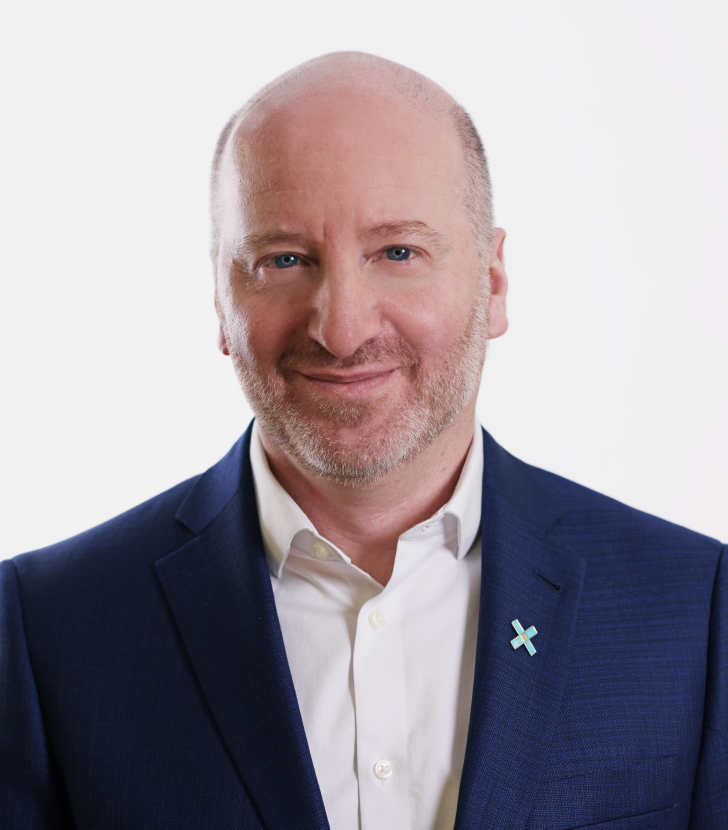 Bald man with a trimmed beard in a blue suit and white shirt, wearing a pin, smiling at the camera against a plain white background.