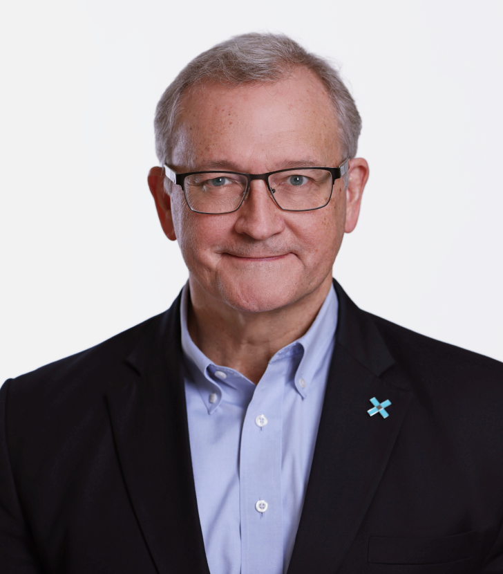 A middle-aged man with gray hair and glasses wearing a dark blazer, light blue shirt, and a pin on his lapel, poses against a plain white background.