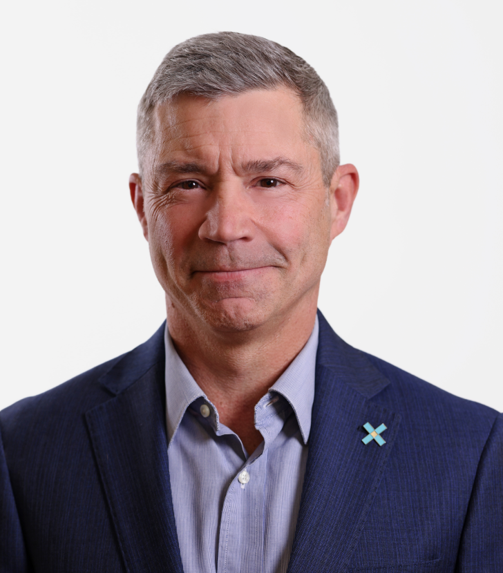 A middle-aged man with short gray hair wears a blue suit jacket and light blue shirt, standing against a plain white background.