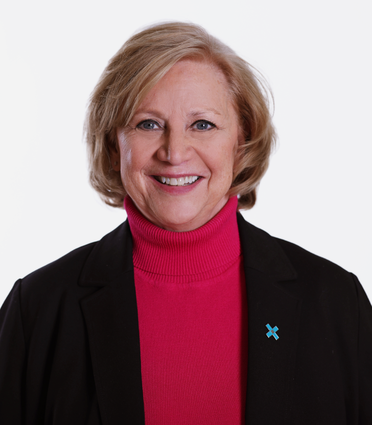 Smiling woman with short blonde hair, wearing a black blazer over a pink turtleneck. A small blue awareness pin is on her left lapel. White background.