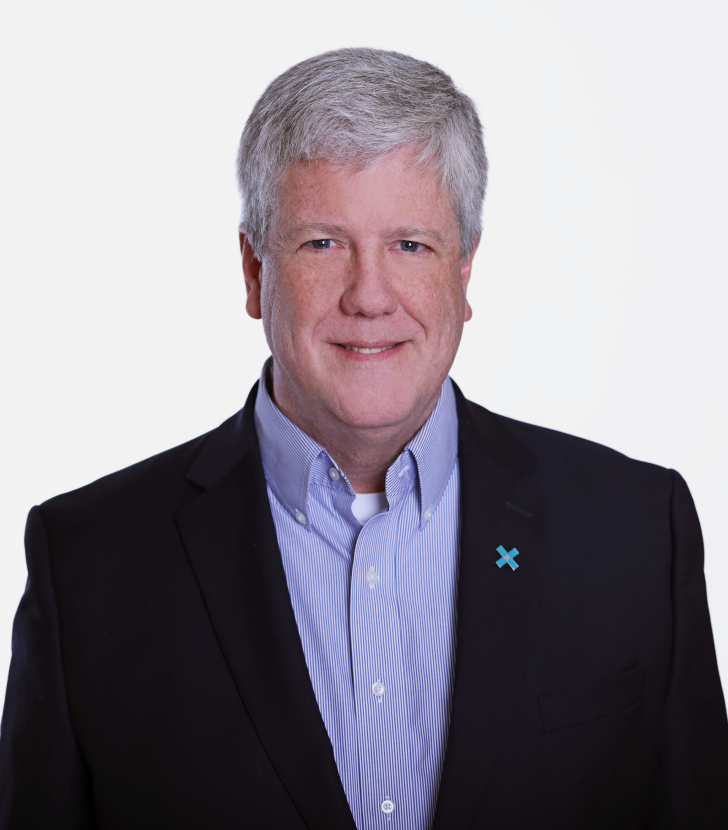 A man with short gray hair wearing a dark blazer, blue shirt, and a pin, smiling at the camera against a plain white background.