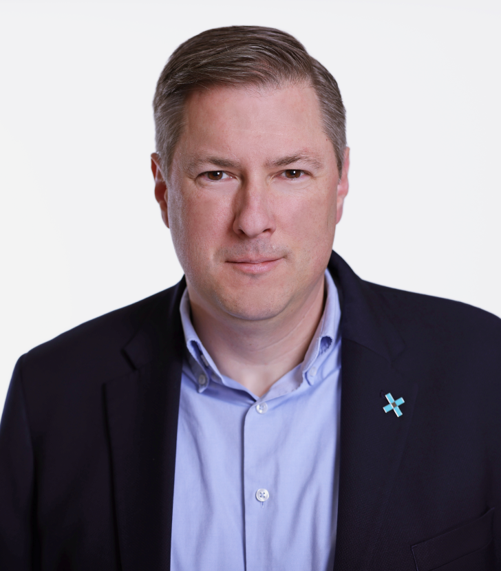 A man with short brown hair wearing a dark blazer, light blue dress shirt, and a blue pin, posing against a plain white background.