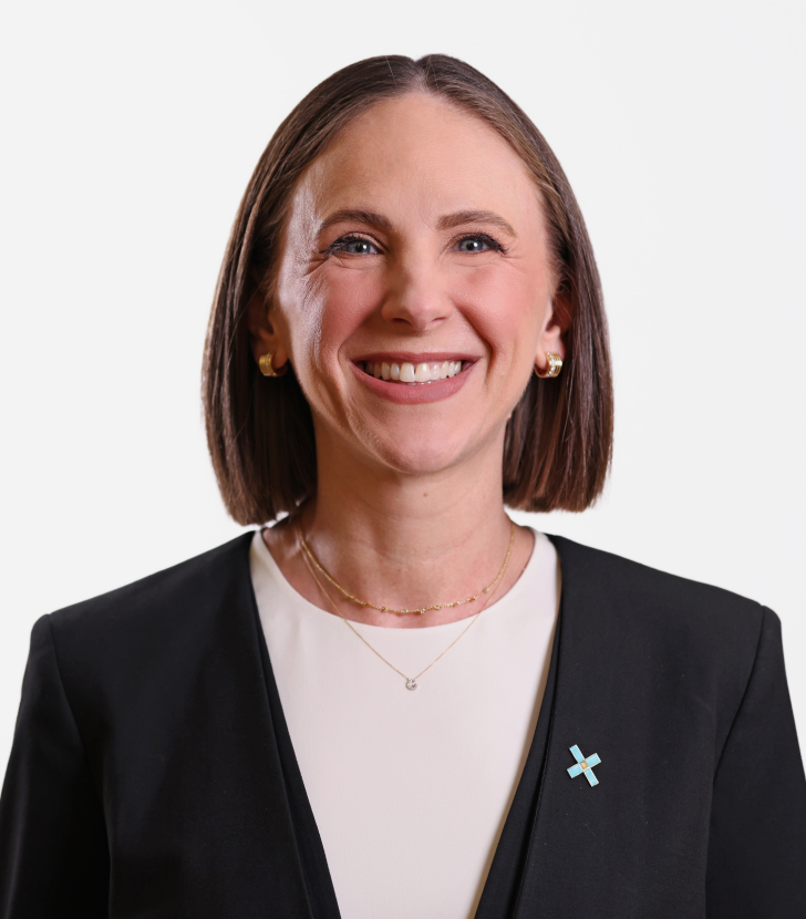 Woman with straight brown hair, wearing a black blazer, white top, and gold jewelry, smiling at the camera against a plain white background.