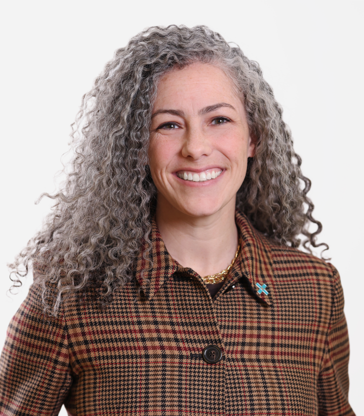 A woman with curly gray hair smiles at the camera, wearing a brown plaid jacket with a blue pin and a gold necklace, against a white background.