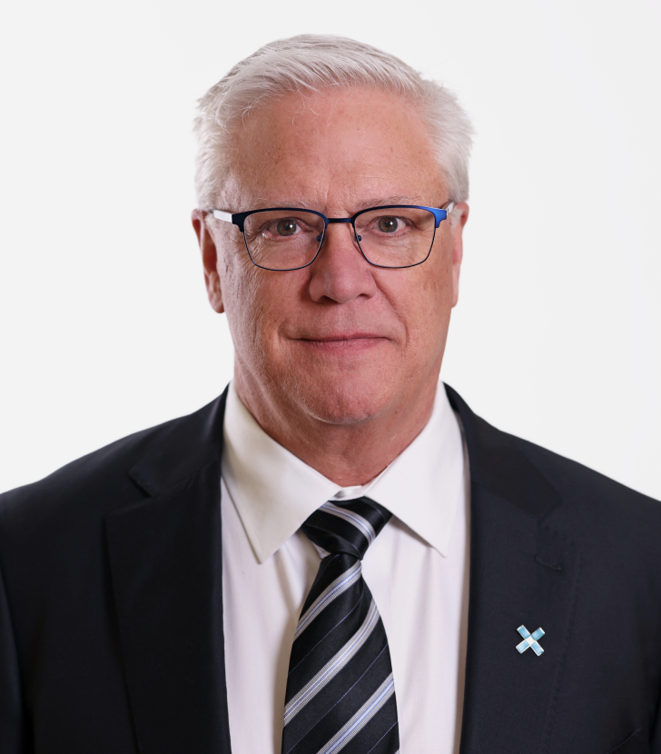 A man with short white hair and glasses wears a dark suit, white shirt, striped tie, and a pin on the jacket, posing against a plain white background.