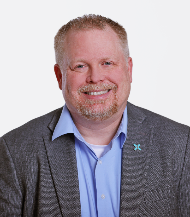 A man with short light hair and a beard, wearing a grey blazer, light blue shirt, and a blue X-shaped pin, smiling in front of a plain white background.