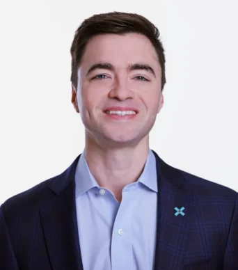 A man with short brown hair wearing a dark plaid suit jacket, light blue shirt, and a pin with an X symbol, smiling at the camera against a white background.