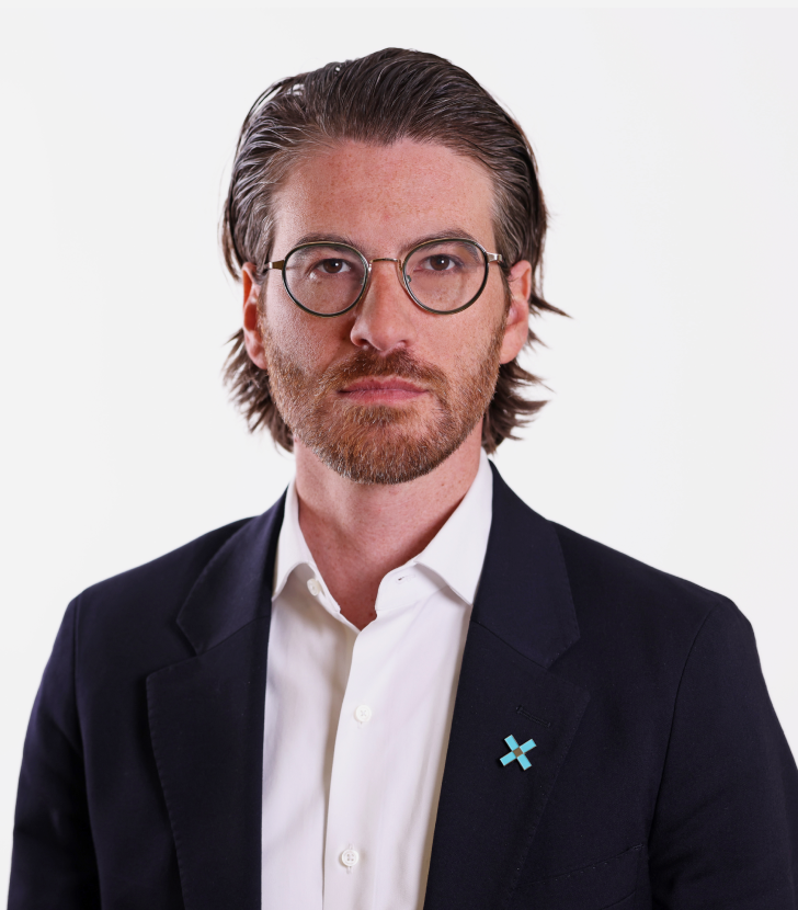 A man with glasses and medium-length brown hair wearing a dark blazer, white shirt, and a blue "X" pin stands against a plain white background.