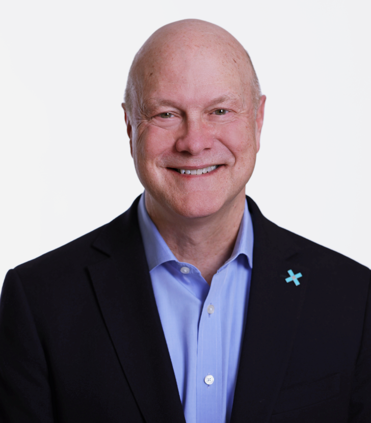 Bald man in a dark blazer and light blue shirt smiling at the camera, with a small blue cross pin on his lapel, against a plain white background.