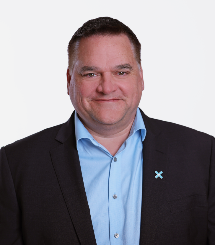 A man in a dark suit and light blue shirt stands against a plain white background, wearing a blue X-shaped pin on his lapel.