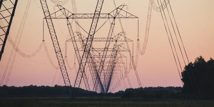 High-voltage power lines and transmission towers stretch across a grassy field at sunset, with trees visible in the background and a pinkish sky overhead.