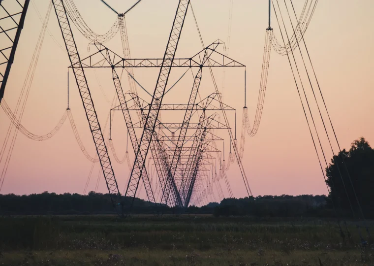 High-voltage power lines and transmission towers stretch across a grassy field at sunset, with trees visible in the background and a pinkish sky overhead.