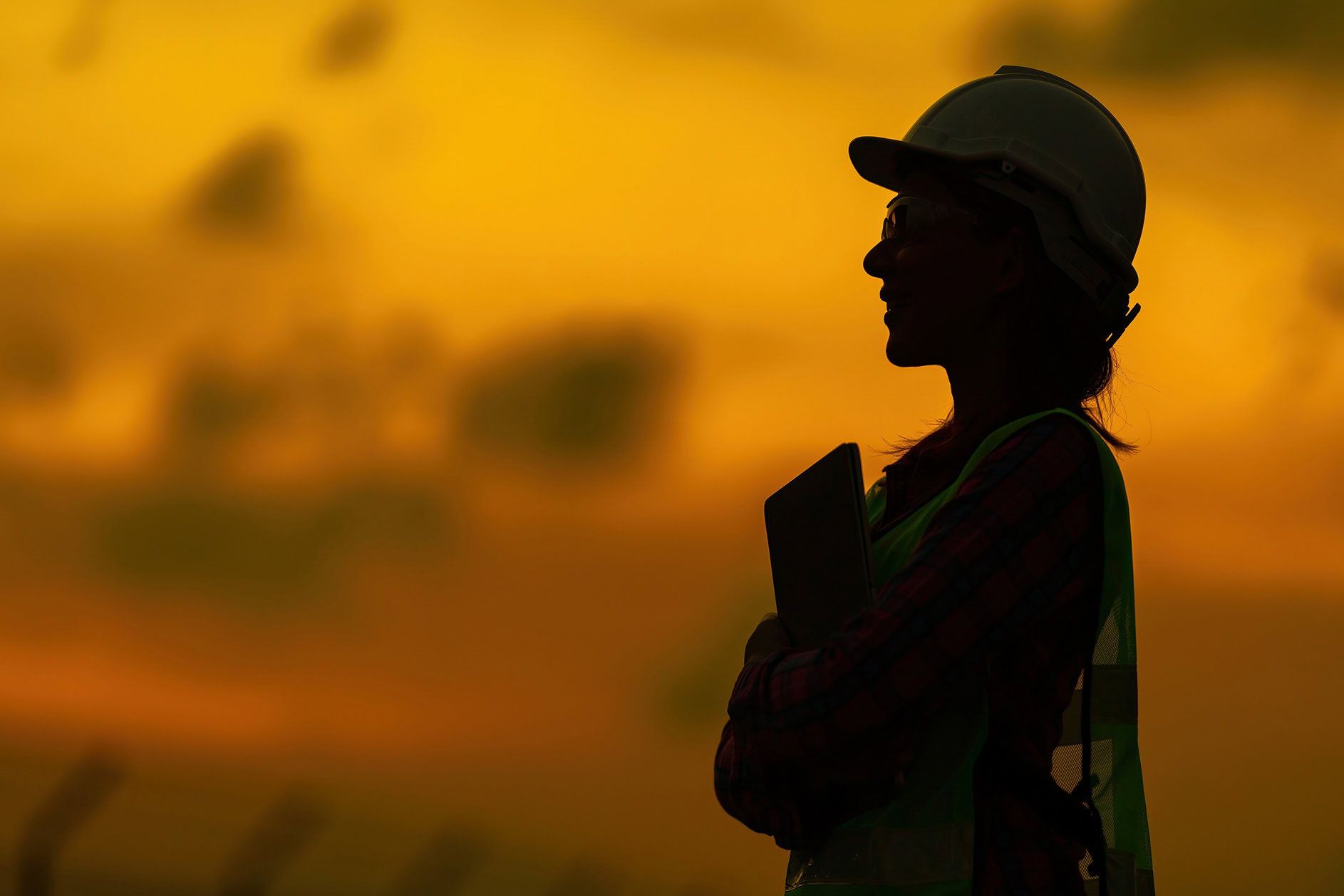 Silhouette of a person wearing a hard hat and safety vest, holding a tablet, standing outdoors at sunset.