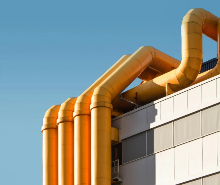 A modern building with several large, yellow industrial pipes running vertically and horizontally against a clear blue sky.