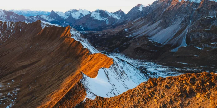 Mountain ridge at sunset with snow patches and distant peaks under a clear sky.