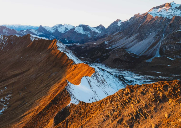 Mountain ridge at sunset with snow patches and distant peaks under a clear sky.