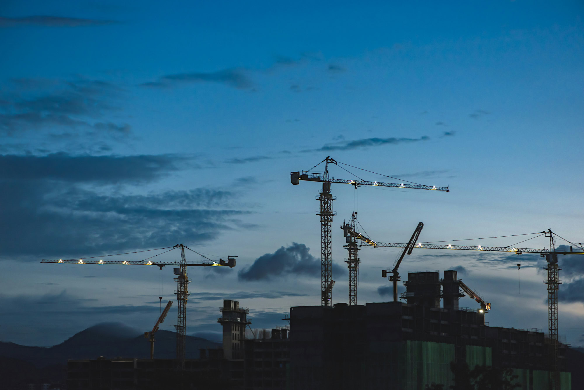 Several tower cranes and unfinished buildings are silhouetted against a blue evening sky with scattered clouds.