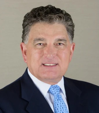 A middle-aged man wearing a dark suit, white shirt, and patterned blue tie poses against a plain beige background.