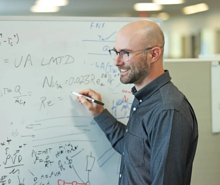 A man with glasses writes equations on a whiteboard in an office setting, smiling while holding a marker.