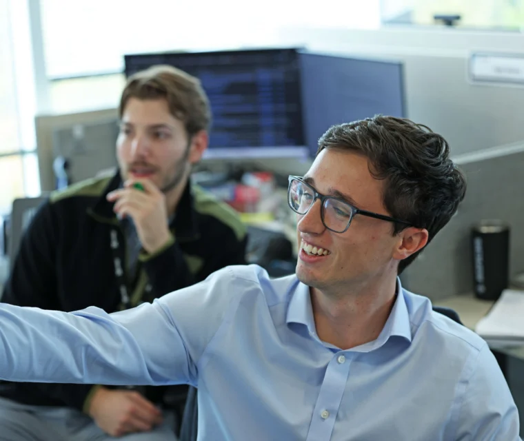 Two people sit at office desks with computer monitors, one in focus smiling and pointing at a screen, the other blurred in the background, with notebooks and a mug on the desk.