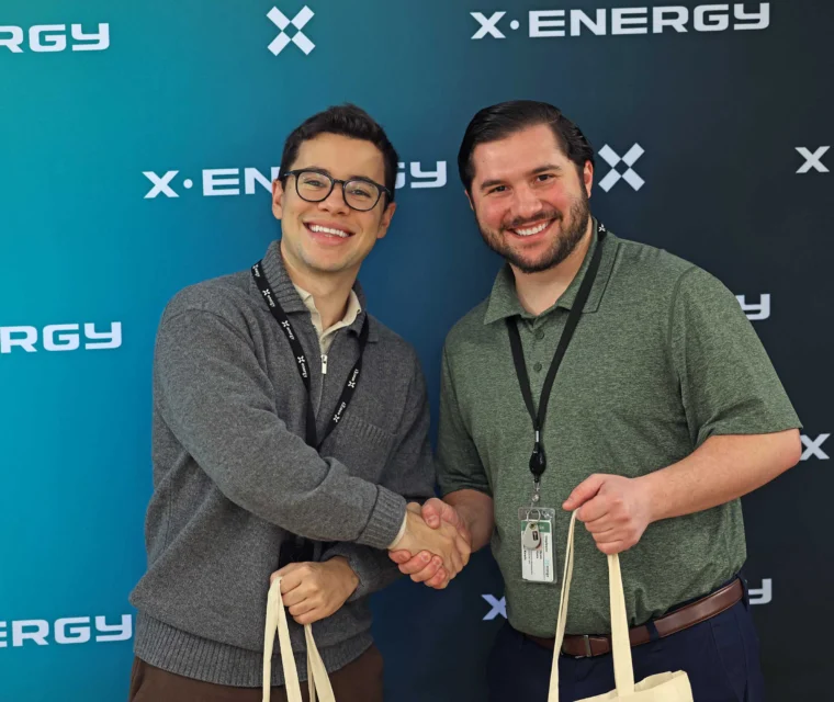 Two men stand in front of an X-Energy backdrop, smiling, shaking hands, and holding tote bags.