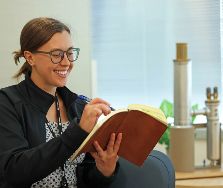 A person wearing glasses and a dark jacket sits indoors, smiling while writing in a brown notebook with a pen.