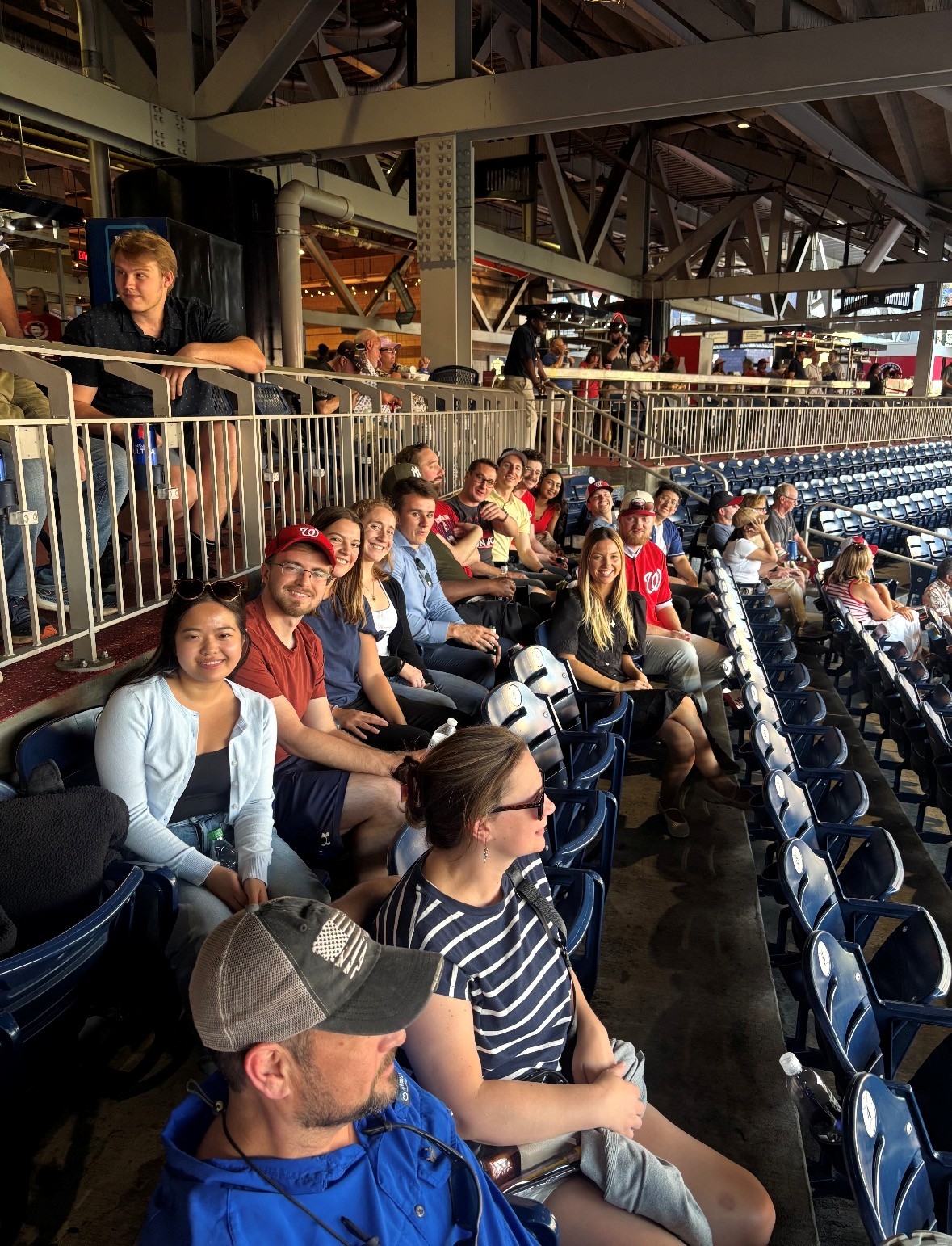 A group of people sit in stadium seats under a covered area, some wearing baseball caps and red shirts, watching a game.