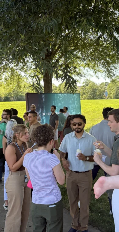 A group of people gather in a shaded area of a park on a sunny day, talking and holding drinks; a baby is held in the foreground.
