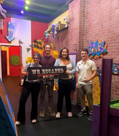 Four people stand in a colorful escape room, smiling and holding a sign that reads “WE ESCAPED.” Bright decorations and toys are visible on the walls and ceiling around them.