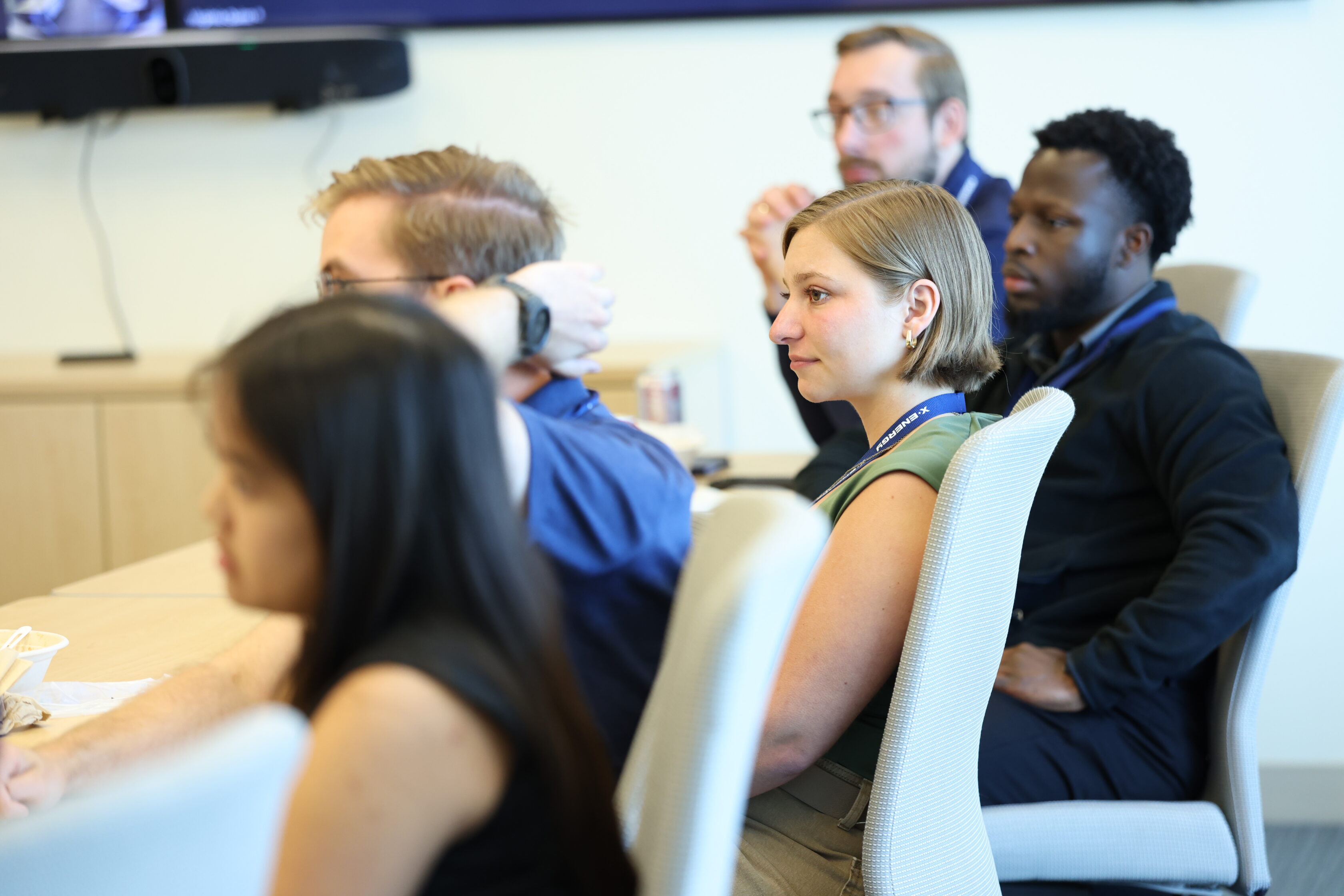 Several people sit attentively at a conference table during a meeting, facing forward, with notepads and lanyards visible.
