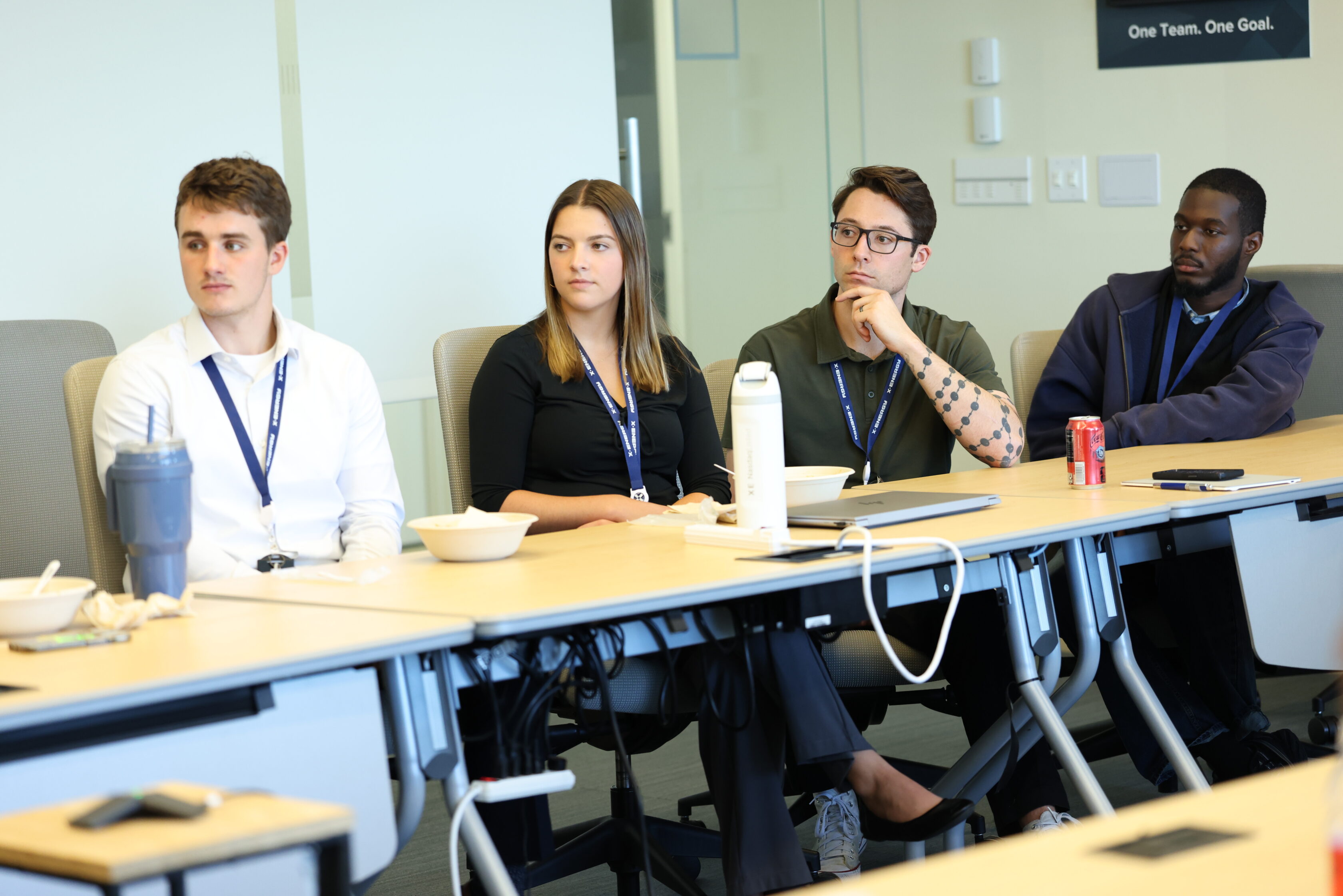 Four people sit at a conference table with drinks and notebooks, attentively listening during a meeting in a modern office setting.