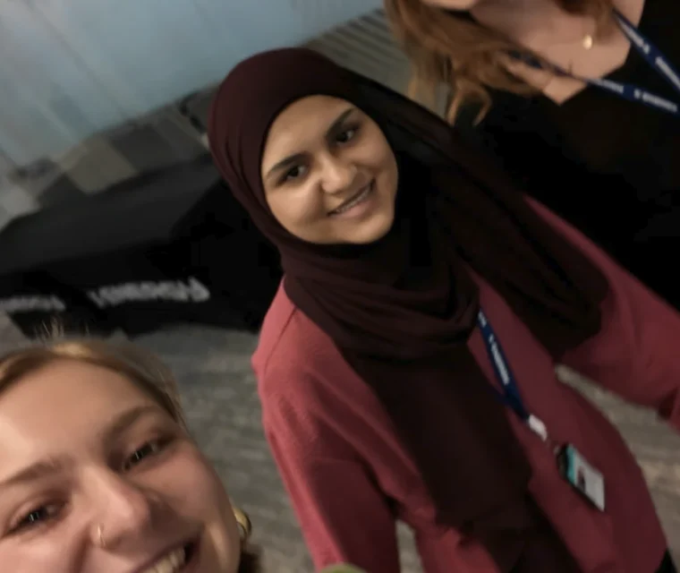 Three women wearing conference badges smile for a selfie indoors, with a blurred background featuring geometric glass walls and tables.