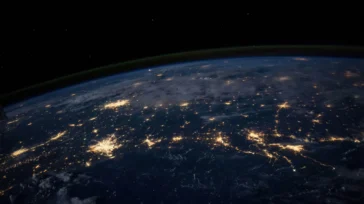 The Earth at night seen from space, with city lights visible across the dark landscape and a faint glow above the horizon.