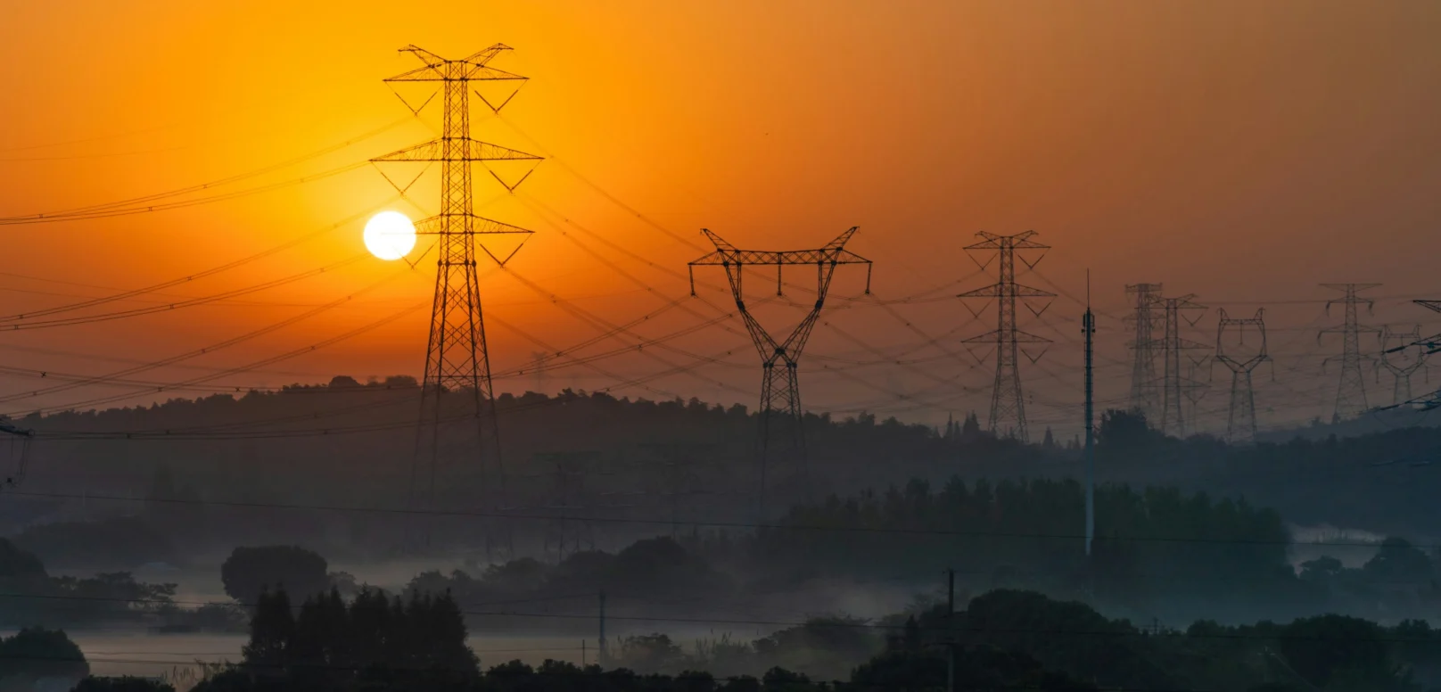 Electricity pylons silhouetted against a sunrise over a hazy landscape.